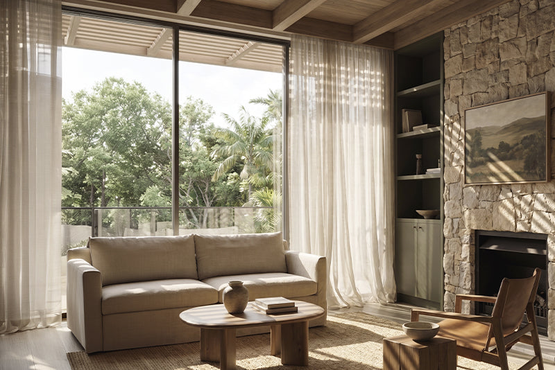 Sunlit living room with sheer curtains beside a stone fireplace.